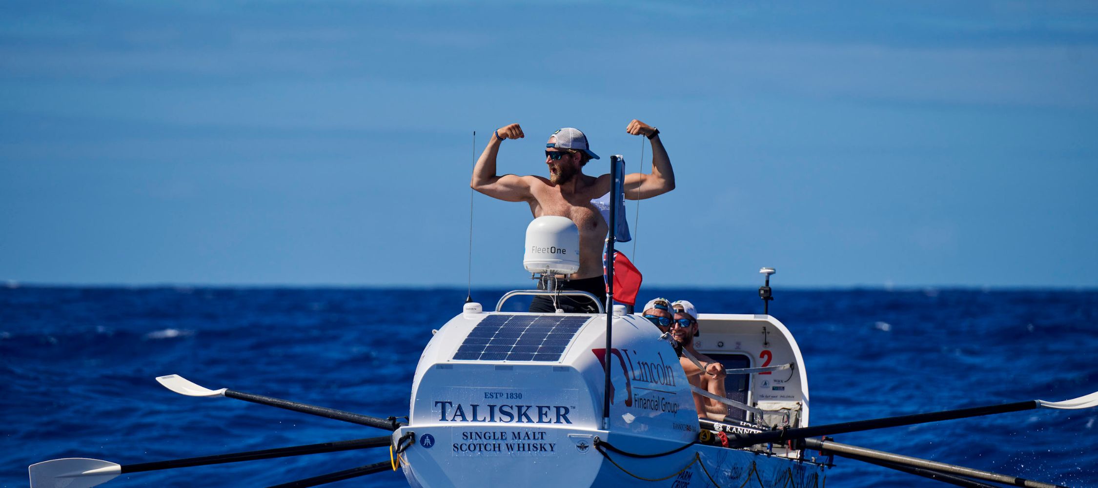 Two men are in a small rowing boat on the ocean. One man, shirtless, flexes his muscles triumphantly while the other sits behind him. The boat is branded with logos and has solar panels on top, with clear blue skies and ocean waters in the background.