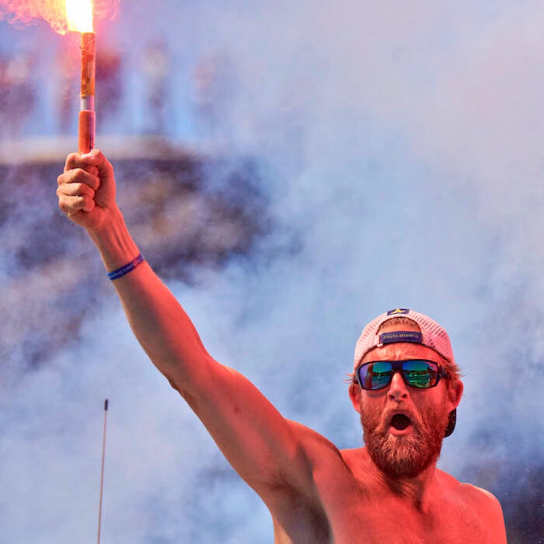 A man with a beard and sunglasses raises a flare above his head, surrounded by smoke. He appears enthusiastic and is shirtless, suggesting a lively sporting event or celebration.