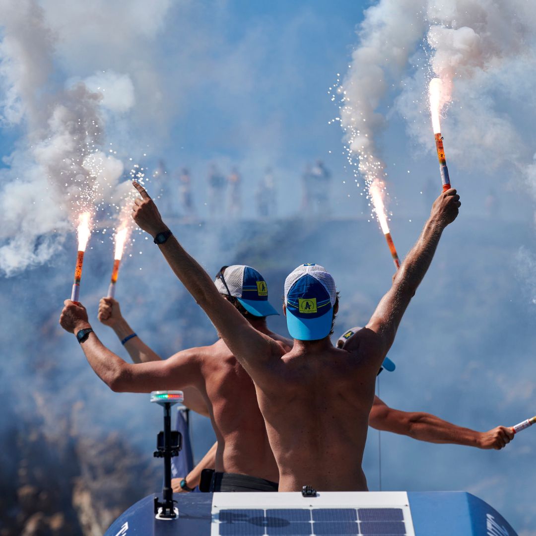 Two shirtless men on a boat raise flares into the air, surrounded by smoke, while a crowd is visible in the background. The scene conveys excitement and celebration.