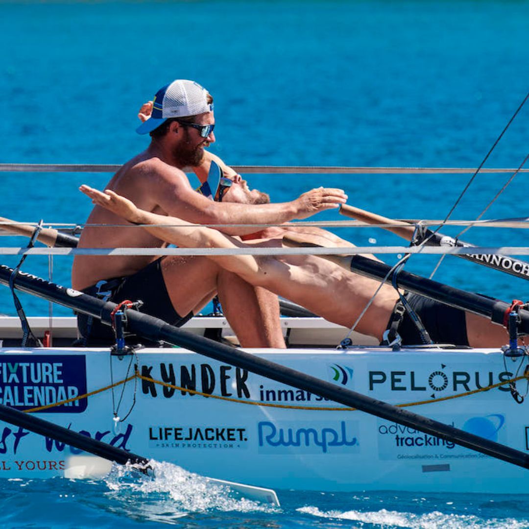 Two rowers in a small boat, intensely focused as they push their oars through bright blue water. One rower is seated at the back, wearing a cap and sunglasses, while the other lies back in a reclined position, both displaying determination in their expressions.