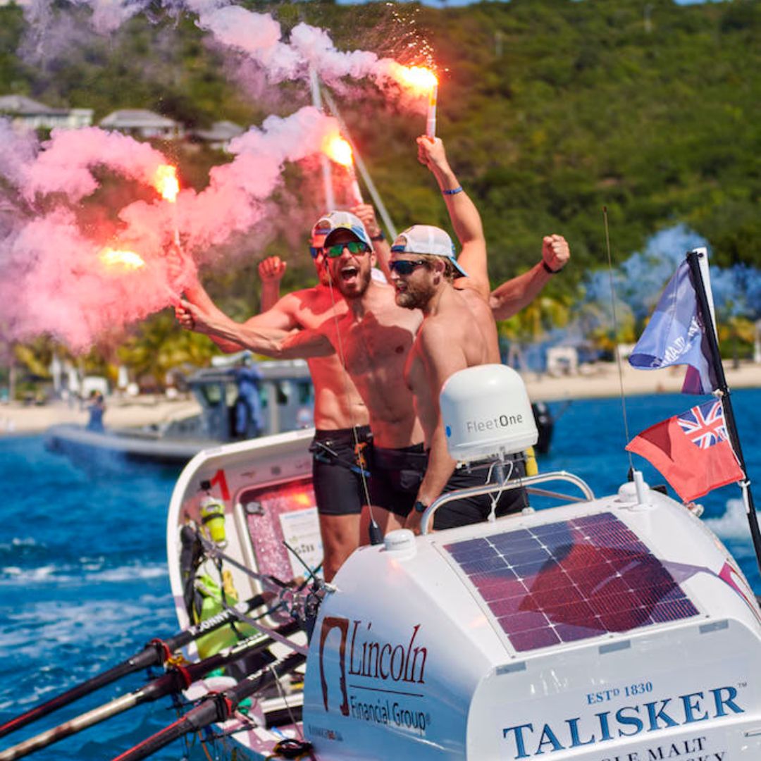 Three athletic men celebrate on a small boat while holding flares. They are shirtless, wearing sunglasses and hats, with a backdrop of water and greenery. Flags are visible on the boat, and there is a vibrant display of smoke from the flares.