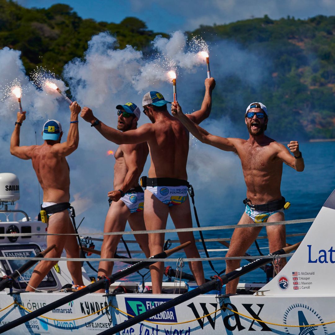 A group of four shirtless men celebrates on a boat, holding flares that emit smoke against a scenic backdrop. They are wearing swim trunks and baseball caps, exuding excitement and camaraderie.