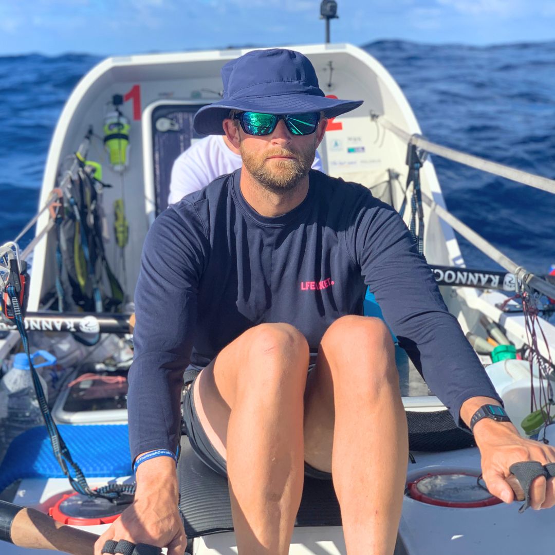 A man with sunglasses and a wide-brimmed hat rows a small boat, wearing a long-sleeve shirt and shorts. The ocean stretches out behind him, with rippling waves and a clear blue sky.