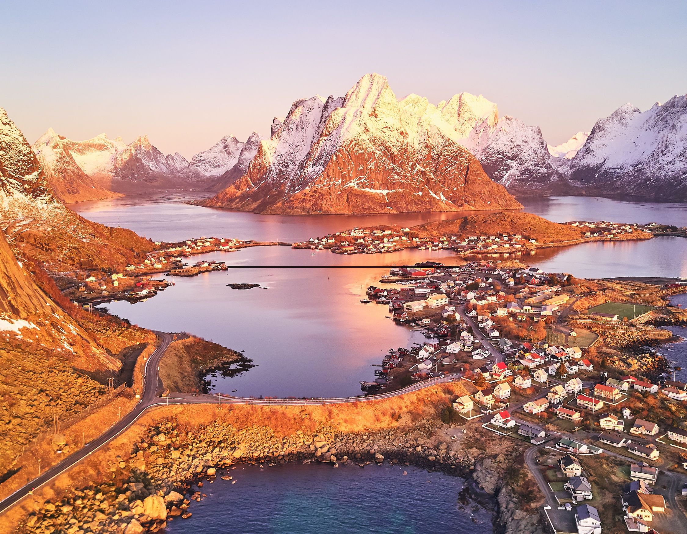 Aerial view of a coastal village with red and white houses nestled between calm water and snow-capped mountains at sunset.