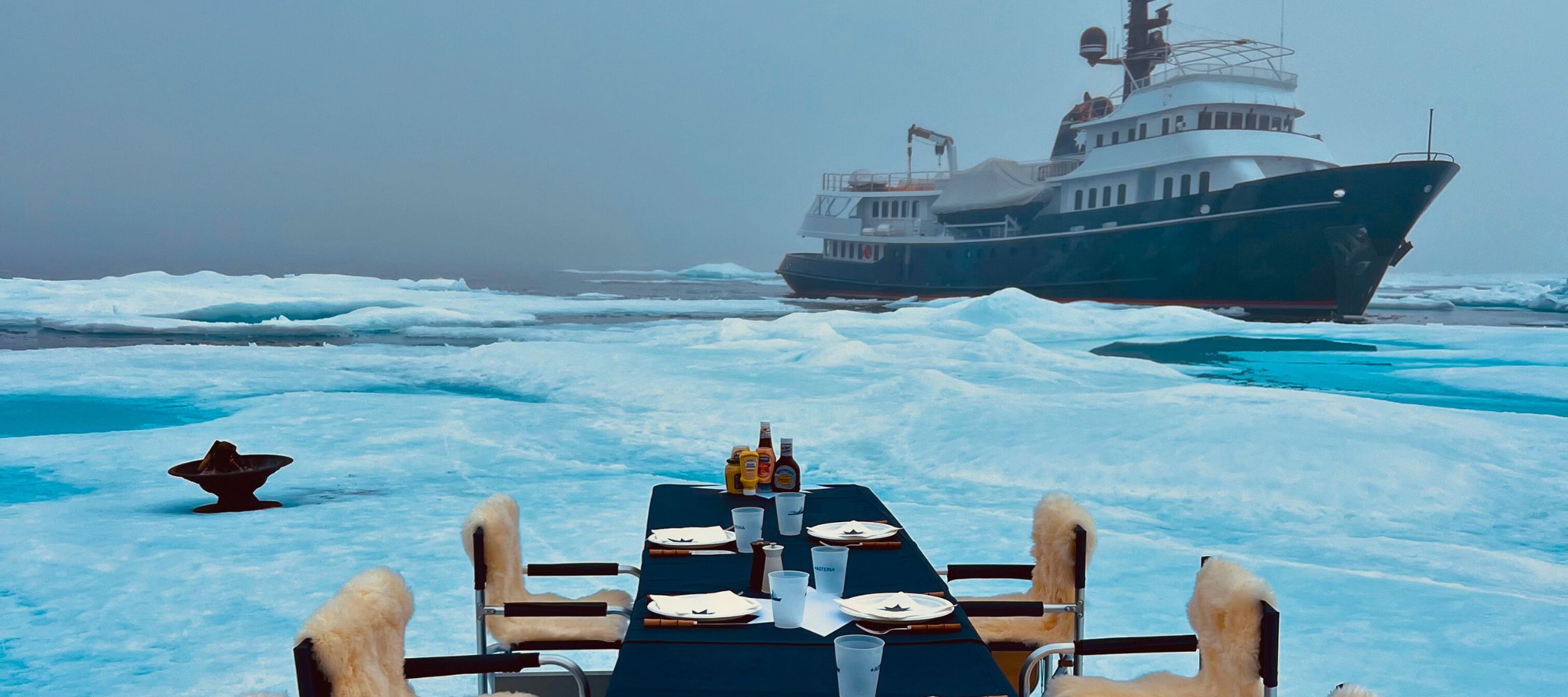 A luxurious dining table set on ice, surrounded by snowy terrain, with a large yacht anchored in the background. The table is elegantly adorned with plates and condiments, creating a surreal dining experience in a cold, arctic environment.