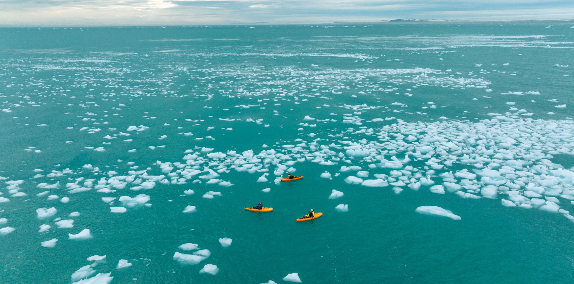Three kayakers in bright orange kayaks navigate through a sea filled with icebergs. The turquoise water contrasts with the white ice, under a cloudy sky.