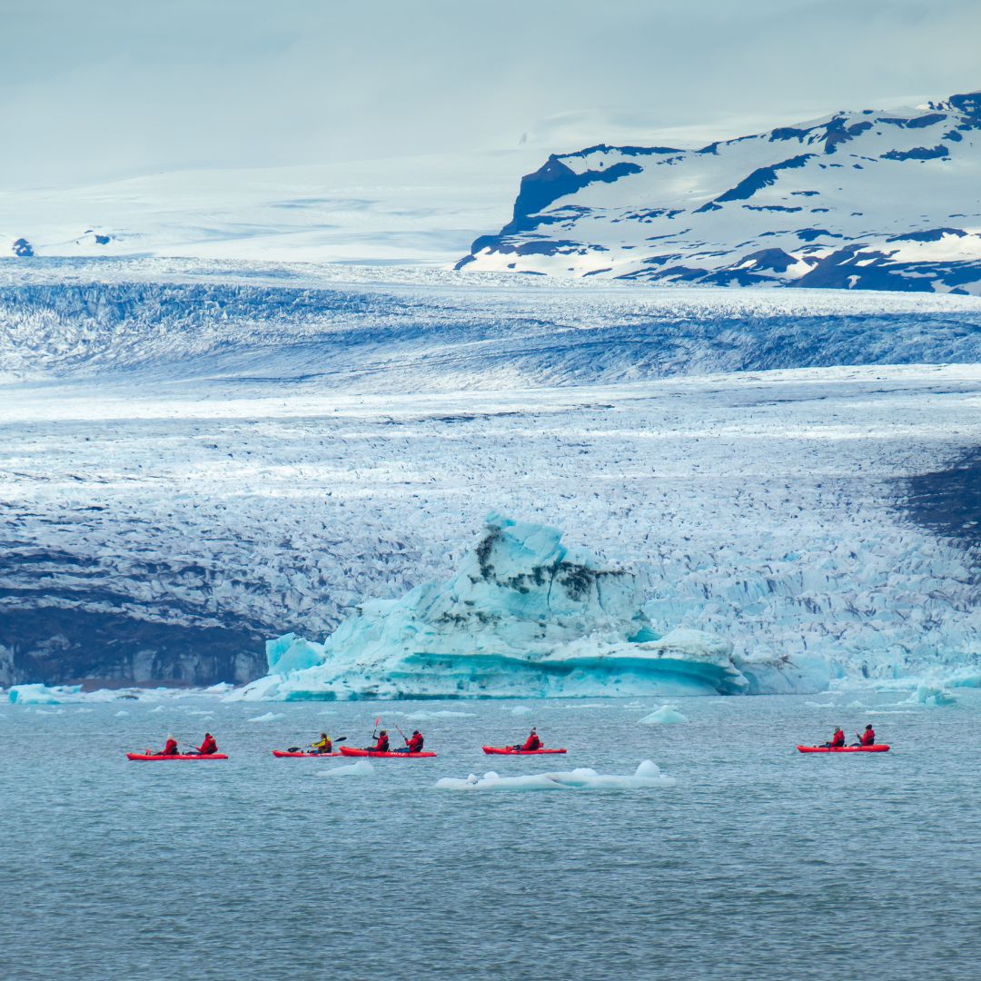 Kayakers in red kayaks paddle across a glacial lagoon, surrounded by icy blue glaciers and snow-capped mountains in the background.