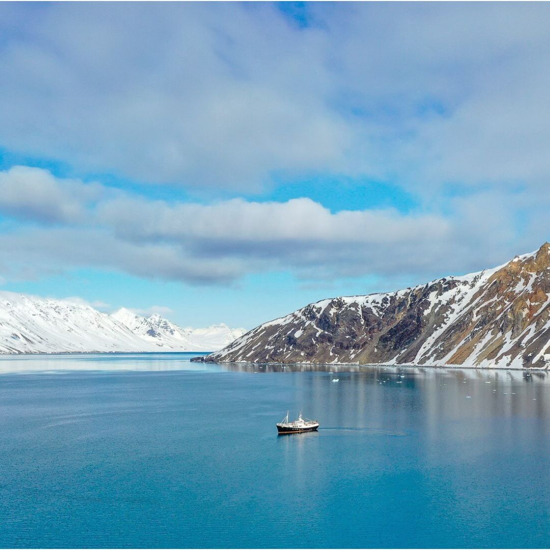 A small ship glides through calm, blue waters surrounded by snow-capped mountains under a partly cloudy sky.