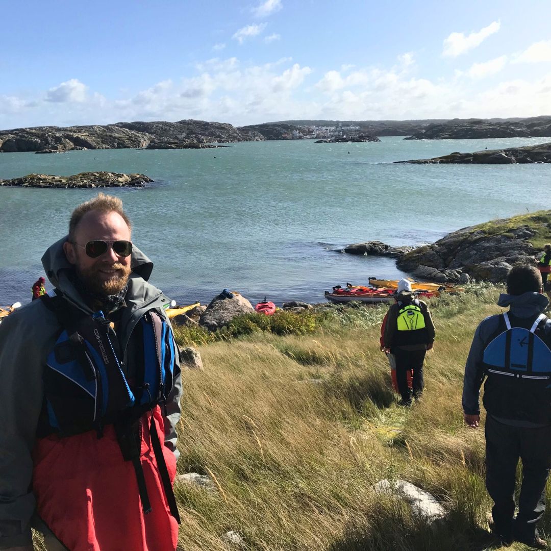 A man in a life jacket stands in tall grass near a rocky shore. Behind him, other people in life jackets walk towards a waterway with several colorful kayaks. The sky is partly cloudy, and hills are visible in the background.