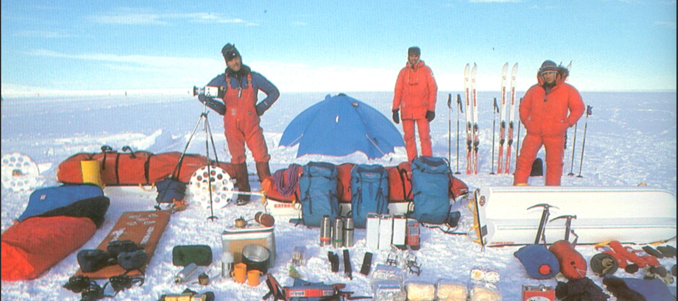 Three individuals in bright red winter gear stand on a snowy landscape, surrounded by various outdoor equipment and supplies. A blue tent is set up in the background, and ski gear is visible on the side. The scene captures an expedition preparation in a cold environment.