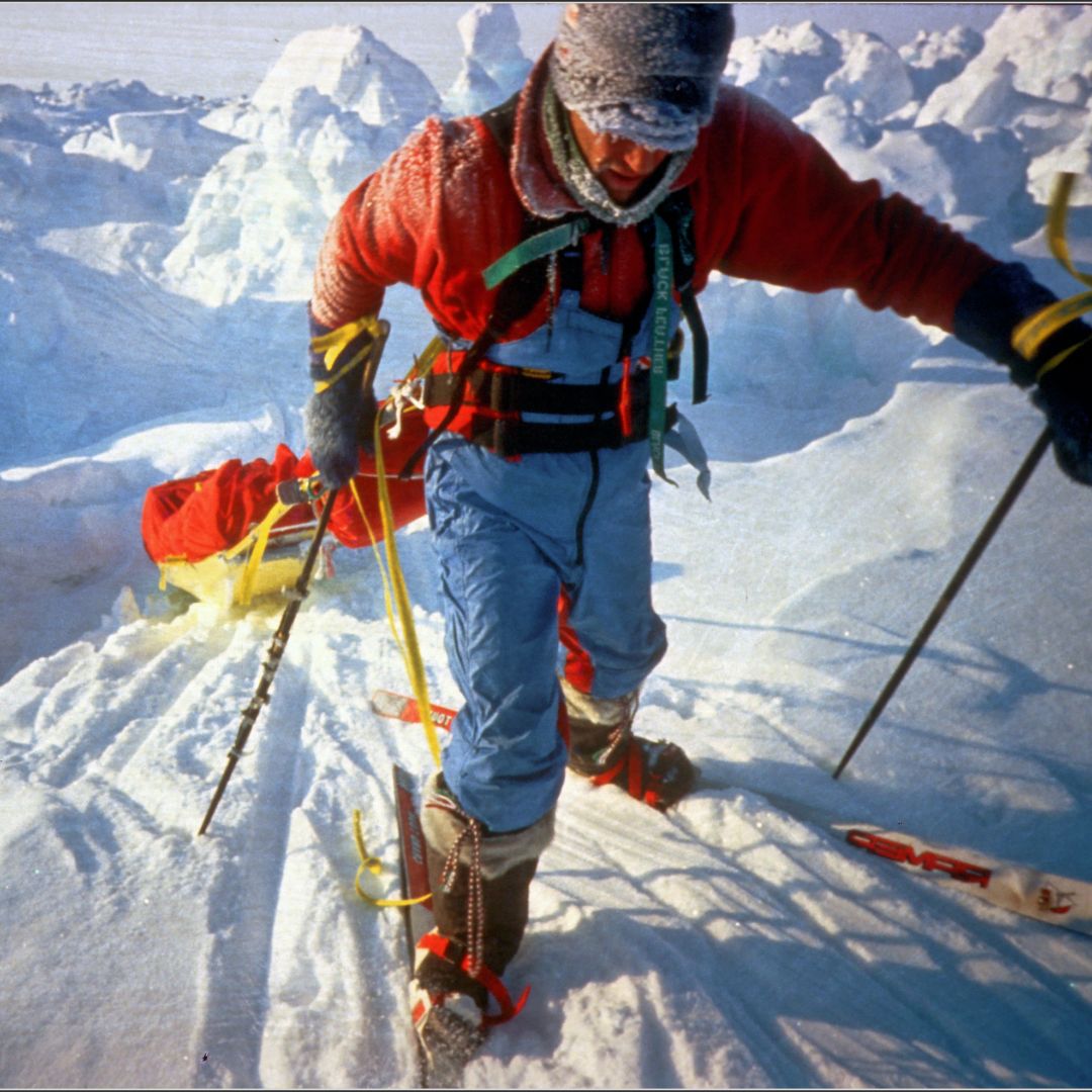 A climber in a red jacket and blue trousers navigates through a snowy, icy landscape. They are equipped with skis, a backpack, and climbing gear, showcasing a rugged outdoor environment.