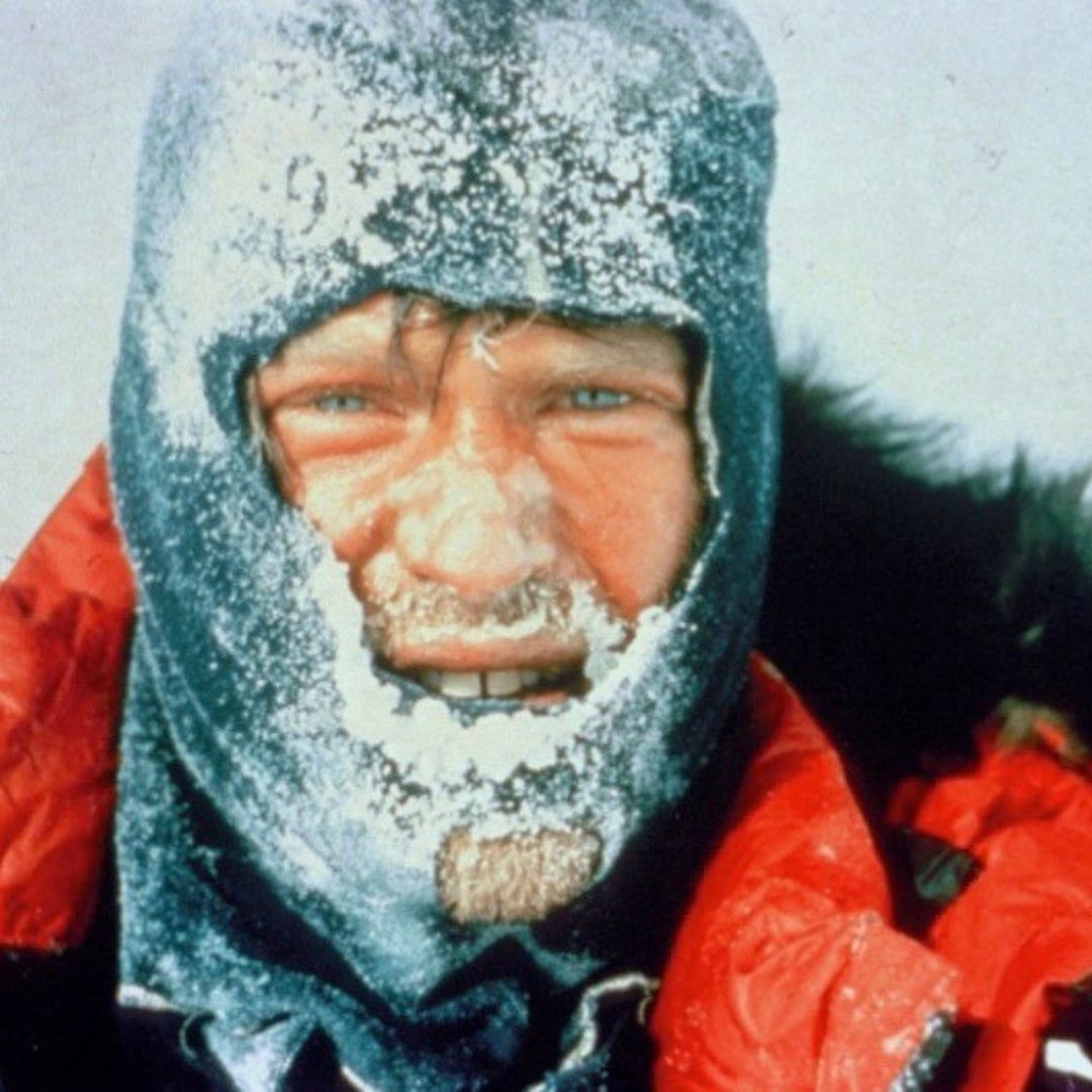 A person in cold weather gear with a snow-covered face and icy eyebrows, smiling slightly. Their red jacket contrasts against the snowy backdrop.