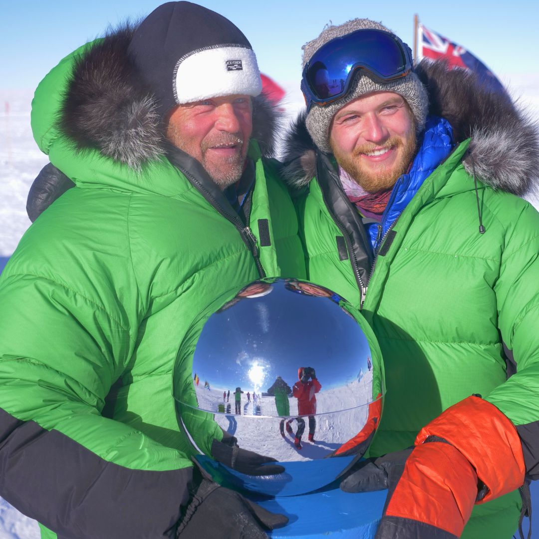 Two men in bright green winter jackets pose together, holding a shiny reflective globe. One man has a beard and is wearing a fur-trimmed hood, while the other has a colorful winter hat and a blue scarf. In the background, blurred figures and flags are visible, indicating a snowy, outdoor environment.