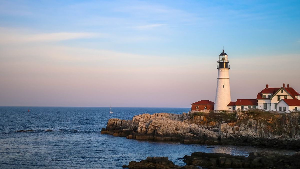 White lighthouse with a black lantern room standing on a rocky cliff beside a red-roofed house at sunset.