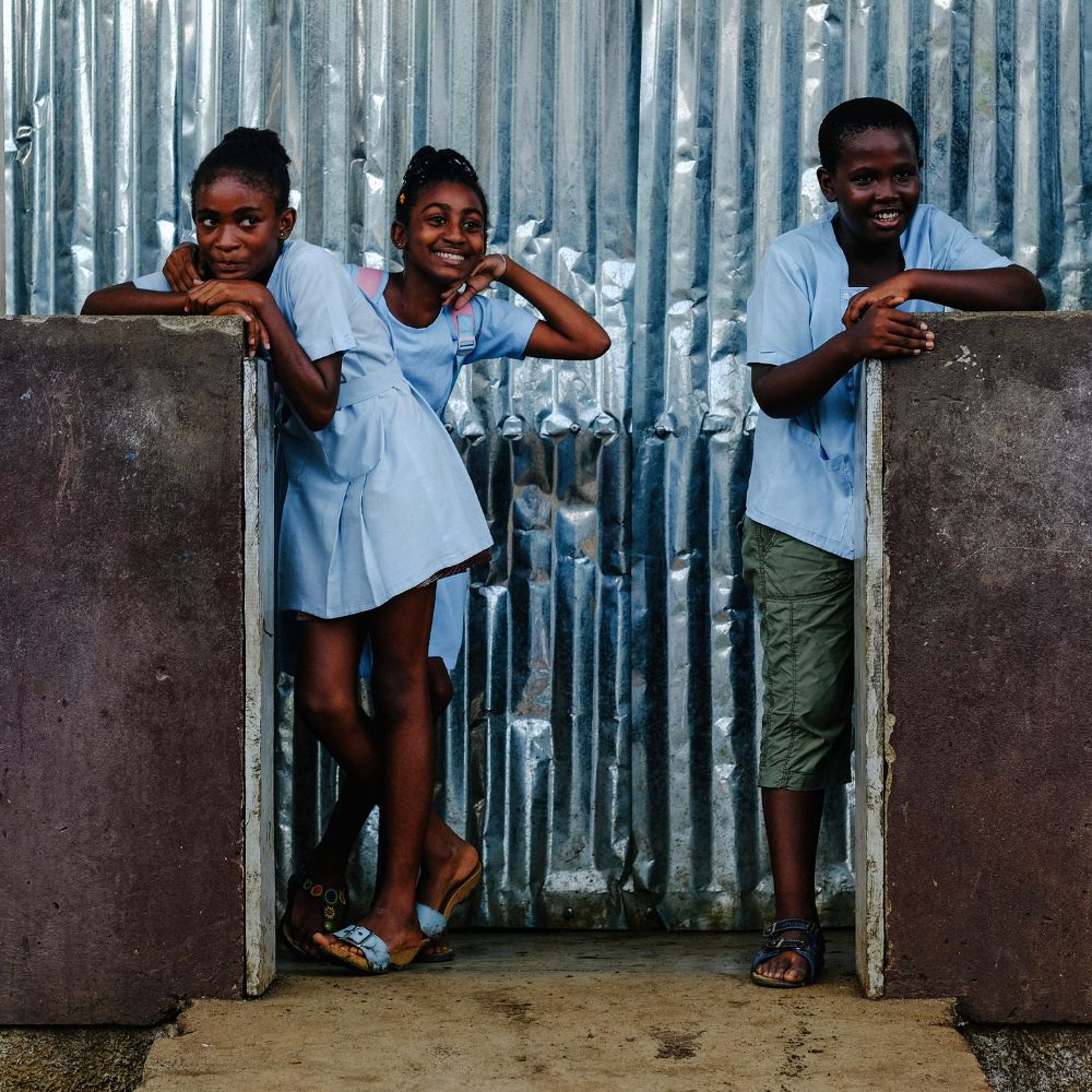 Three children wearing light blue school uniforms lean playfully against a textured, corrugated metal backdrop. Two girls stand with arms resting on a stone wall, while a boy stands beside them, smiling.