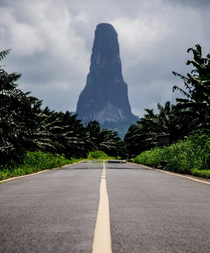 A narrow road stretches towards a towering, jagged peak in the distance, surrounded by lush green vegetation and palm trees under a cloudy sky.