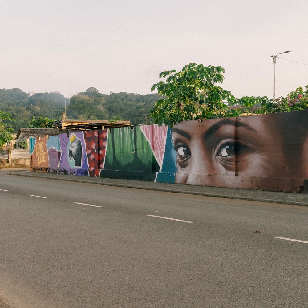 A vibrant mural featuring large, expressive eyes on a wall, surrounded by colorful depictions of fruits and abstract designs. Lush greenery and distant hills in the background, alongside a paved road.