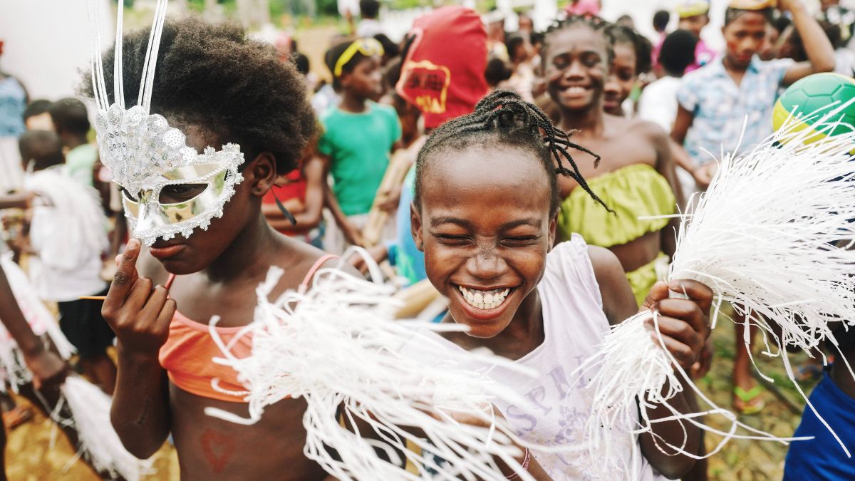Children celebrate at a lively festival, wearing colorful costumes and accessories. One child dons a glittering mask while another joyfully holds white pom-poms, all surrounded by others enjoying the festivities. Bright atmosphere filled with laughter and vibrant clothing.