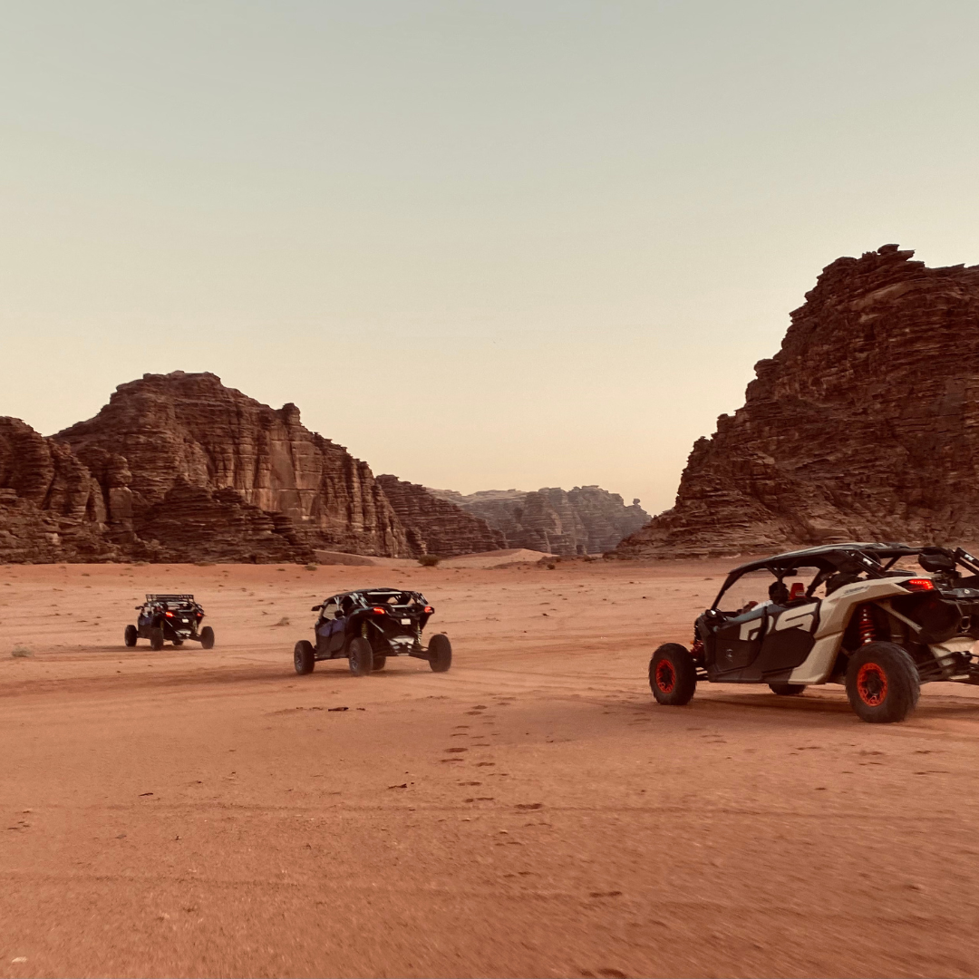 Three off-road buggies drive through a vast desert valley between towering, layered rock formations.