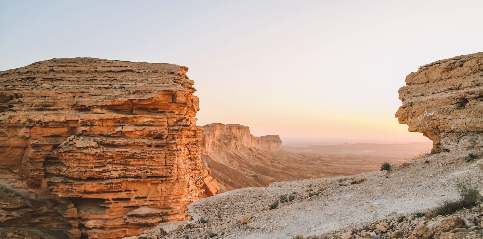 A scenic view of rugged rock formations in a desert landscape during sunset, with layers of orange and beige cliffs illuminated by soft light.
