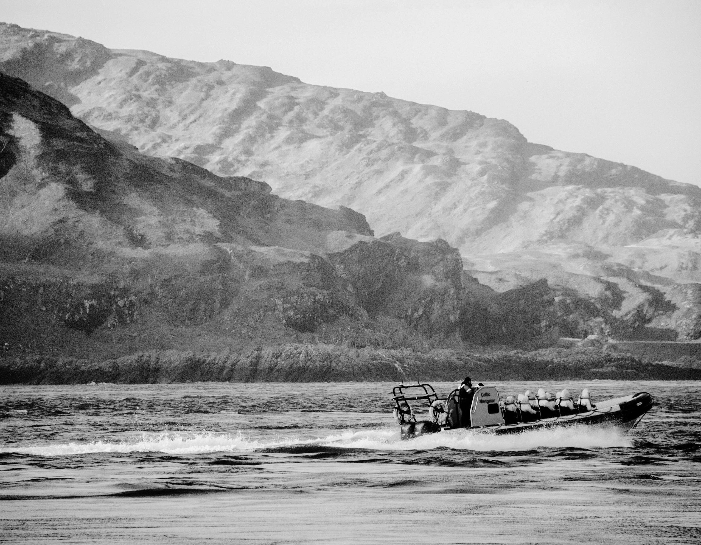 Black and white photo of a motorized raft carrying passengers across choppy water in front of a mountainous shoreline.