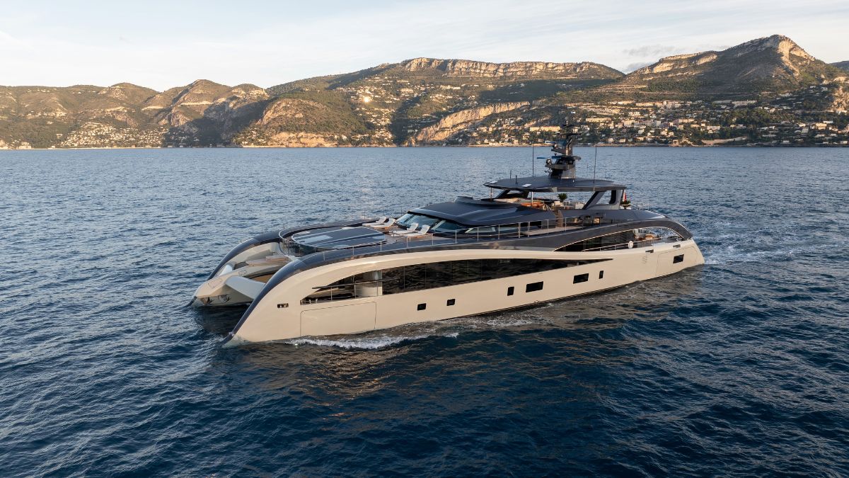 Sleek, tan and black modern catamaran yacht cruising on calm blue water with coastal mountains in the background.