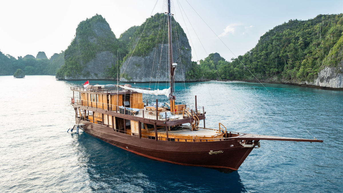 Traditional wooden Phinisi boat with multiple decks anchored in a tropical bay surrounded by lush limestone islands.