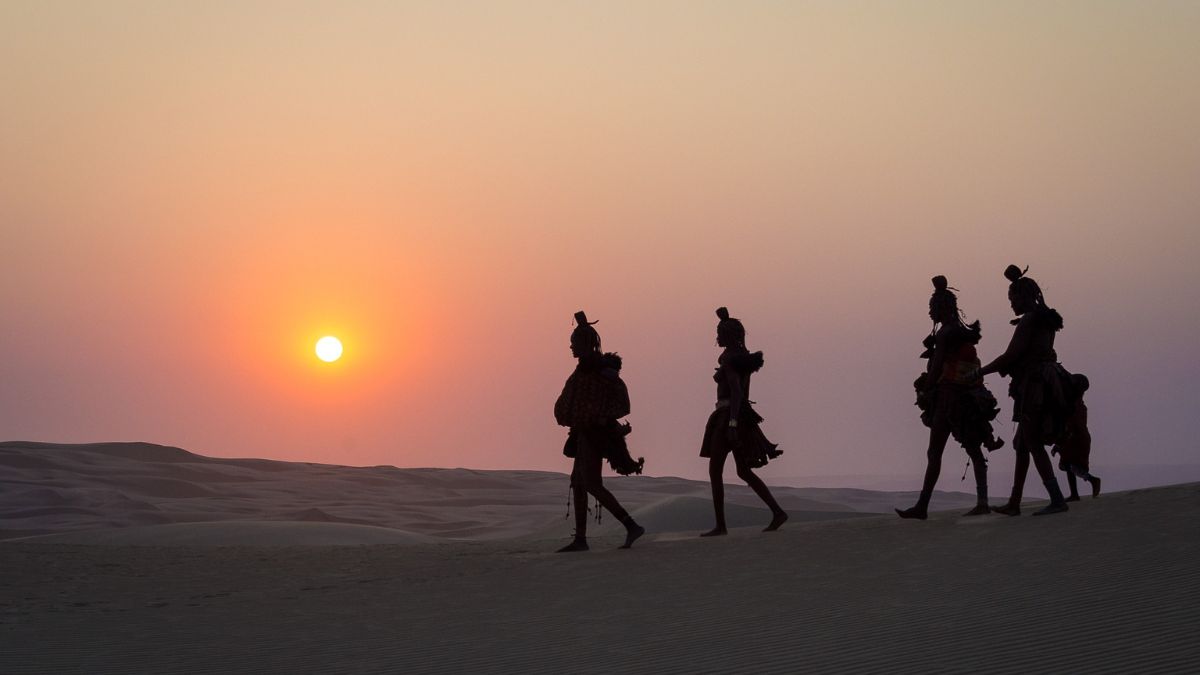 Silhouettes of people walking across desert sand dunes against a hazy orange sunset.