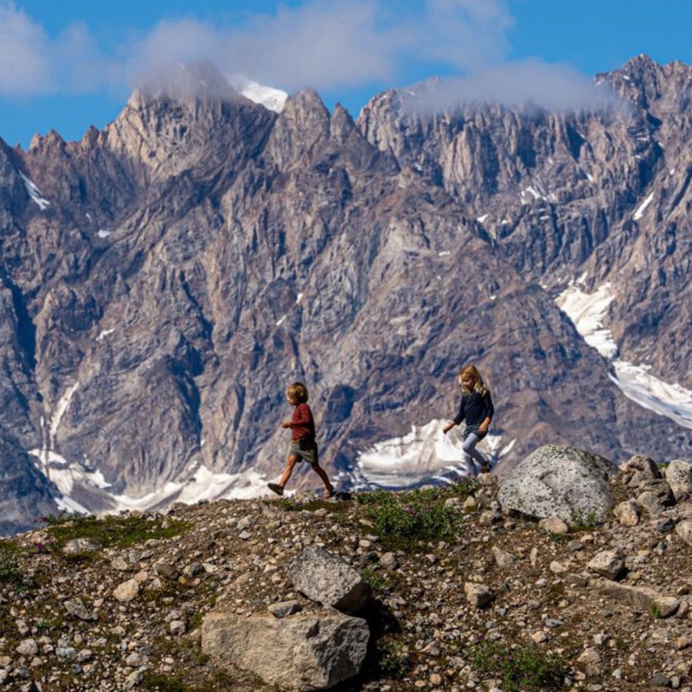 Two children walking on a rocky terrain with majestic mountains in the background, showcasing rugged peaks partially covered by clouds.