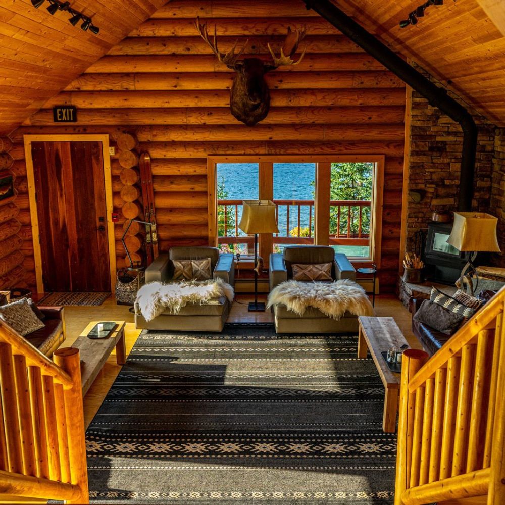 A cozy cabin interior featuring wooden walls and beams, with two stylish armchairs draped in fur, a coffee table, and a patterned rug. Large windows reveal a scenic view of water and trees outside. A mounted deer head hangs above the fireplace.