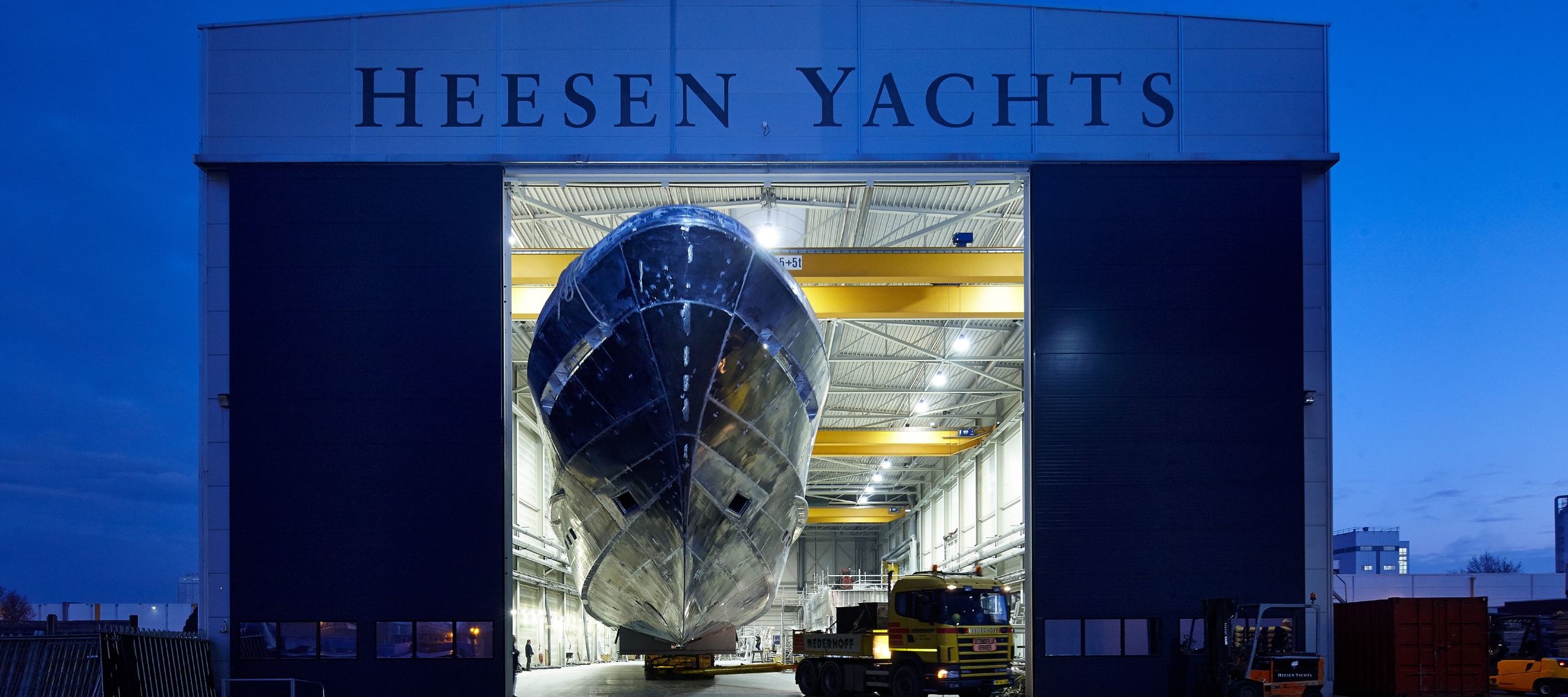 A large, sleek yacht is being moved out of a hangar at Heesen Yachts during twilight, showcasing its polished hull and modern design. Cranes and machinery are visible inside the facility.