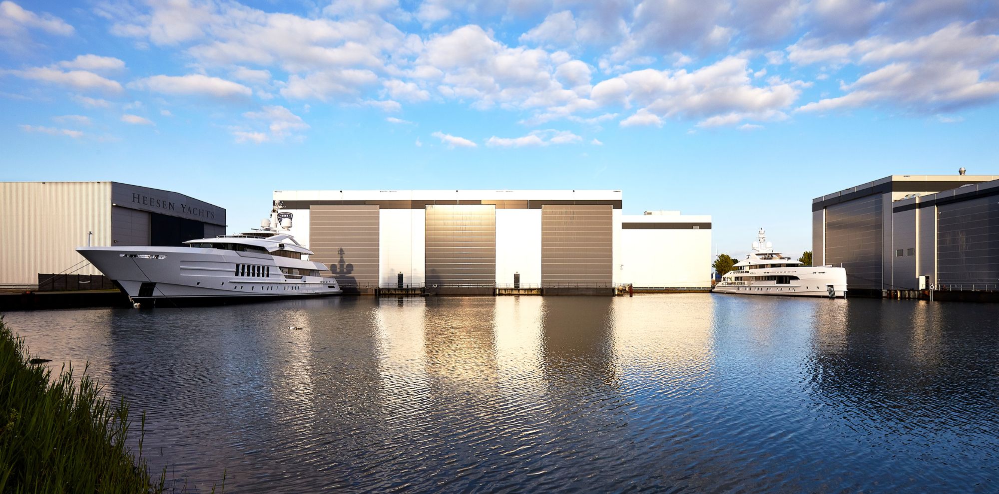 Two large yachts docked at a marina with modern gray storage buildings in the background. The water reflects the blue sky and clouds, creating a serene atmosphere.