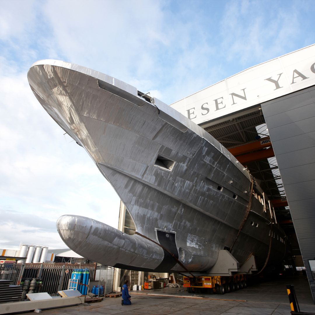 A large, sleek yacht made of polished metal is partially emerging from a shipbuilding facility. The yacht has a modern design with a prominent bow, and workers are visible near the base. Cloudy skies can be seen in the background.