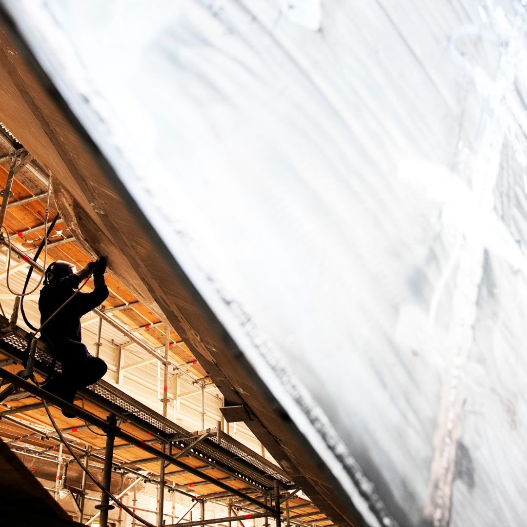 A silhouette of a worker on scaffolding, working on an elevated structure in a large, well-lit industrial space.