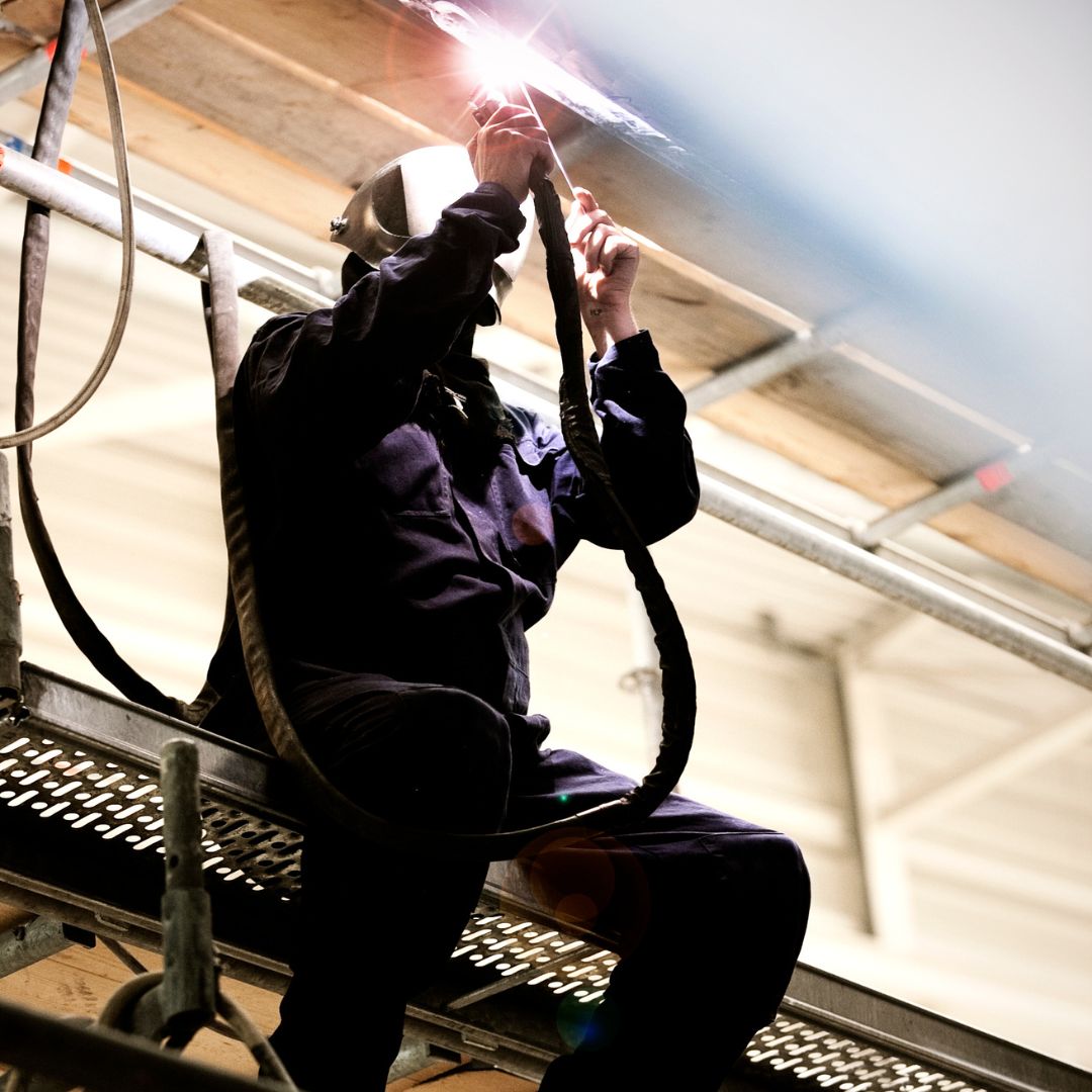 A welder in protective gear works on a metal structure while standing on scaffolding. The bright sparks from the welding process illuminate the scene.