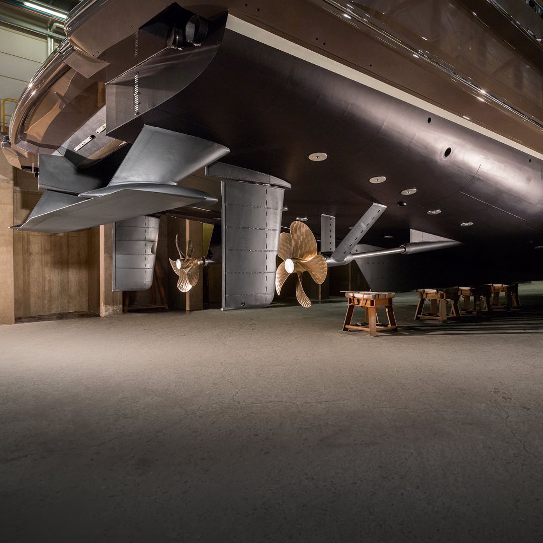 A close-up view of a large yacht's stern showcasing its twin propellers and rudders. The sleek hull is partially illuminated, revealing the vessel's advanced design and craftsmanship. The surrounding area has a concrete floor and wooden stands supporting the boat.