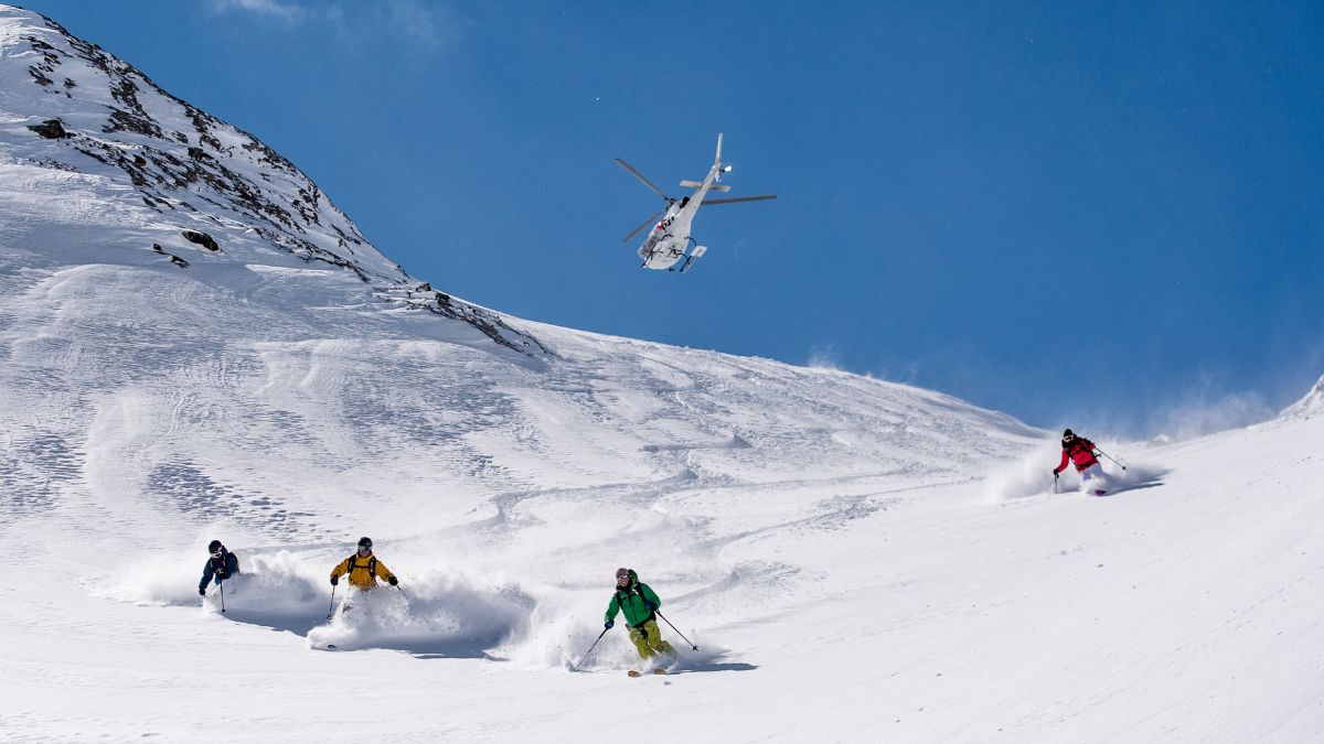 Four skiers descend a steep, snow-covered mountain slope while a white helicopter flies overhead against a blue sky.