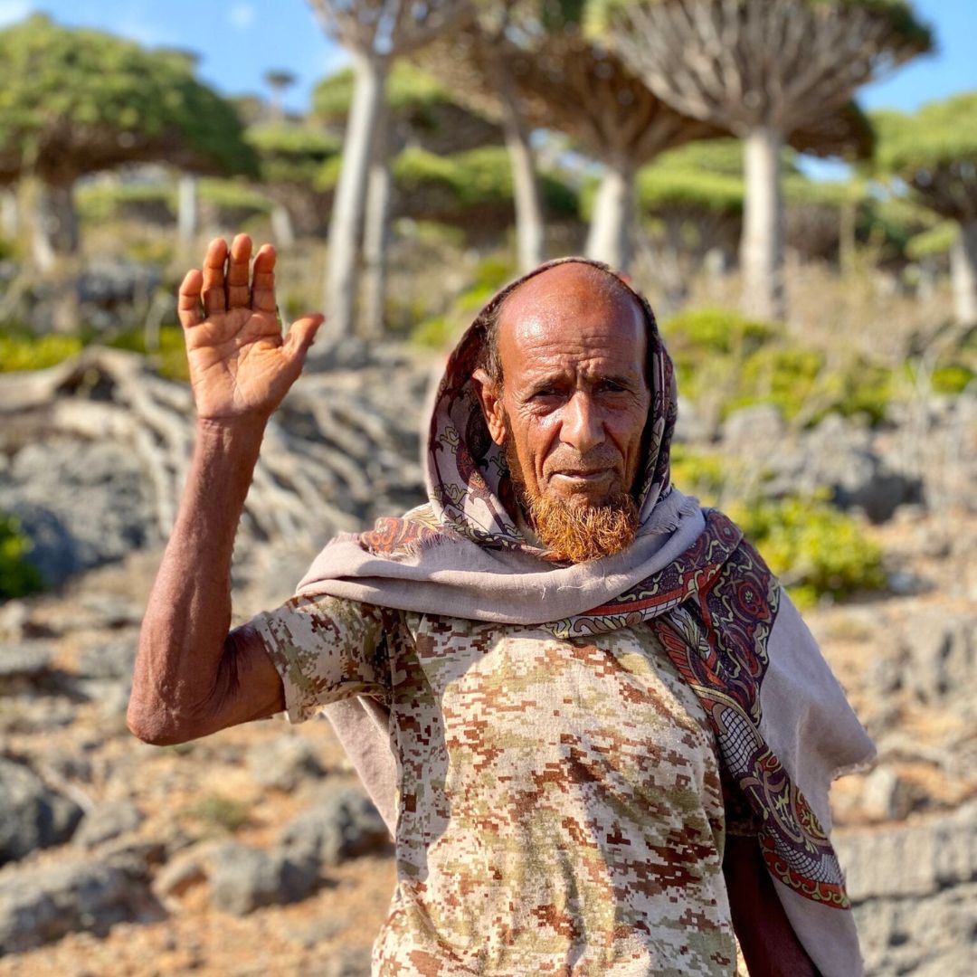 An elderly man with a beard and headscarf raises his hand in greeting, standing on rocky terrain with unique trees in the background.
