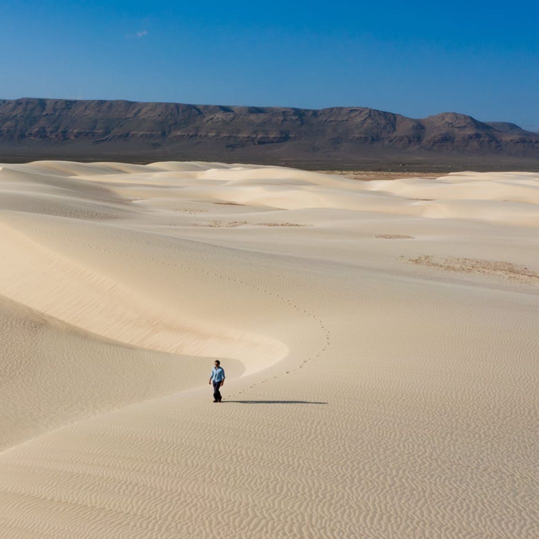 A lone figure stands in an expansive desert landscape, surrounded by rolling sand dunes under a clear blue sky. Gentle shadows create texture across the dunes, while a distant mountain range looms in the background.