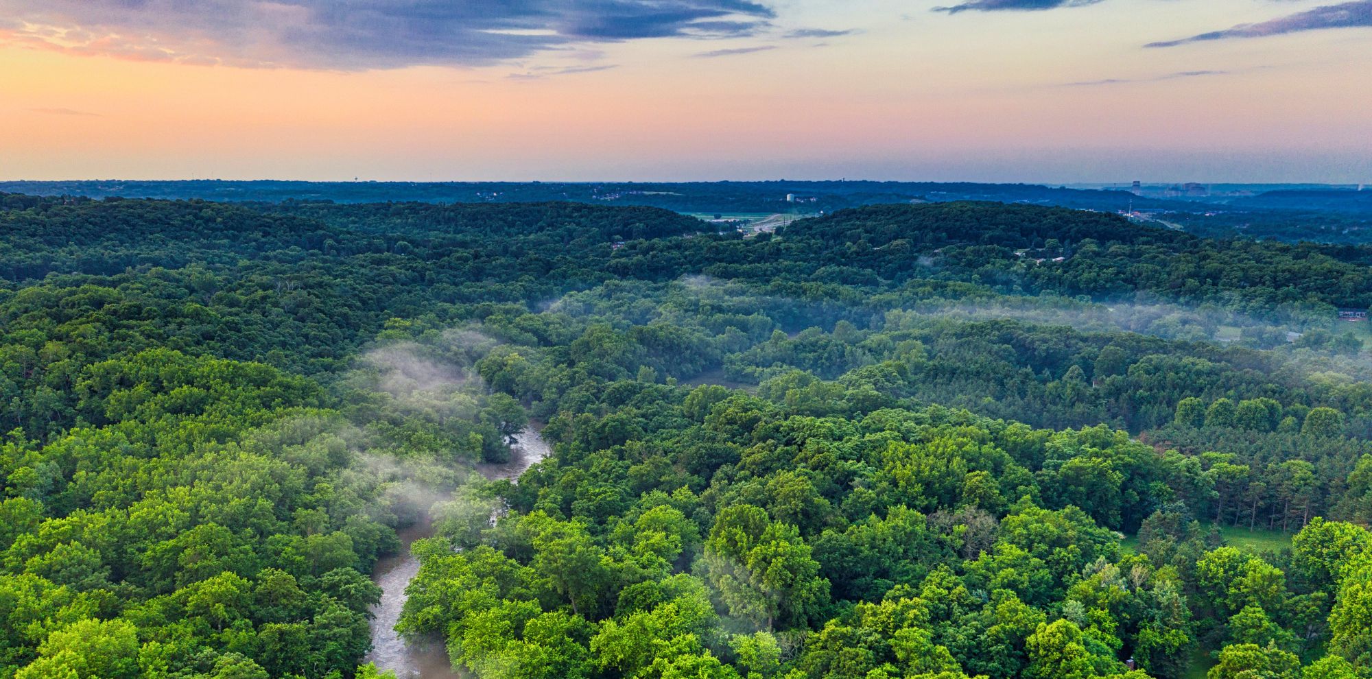 Aerial view of a winding river flowing through a dense, lush green forest with morning mist under a sunset sky.