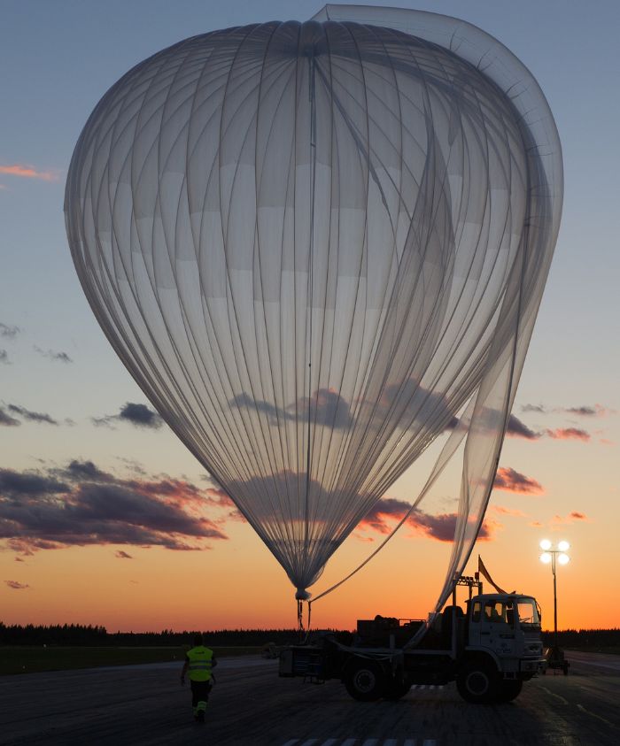 A large, translucent balloon being prepared for launch at sunset, with a silhouette of a person in a reflective vest beside a truck. The sky is filled with soft clouds and hues of orange and purple.