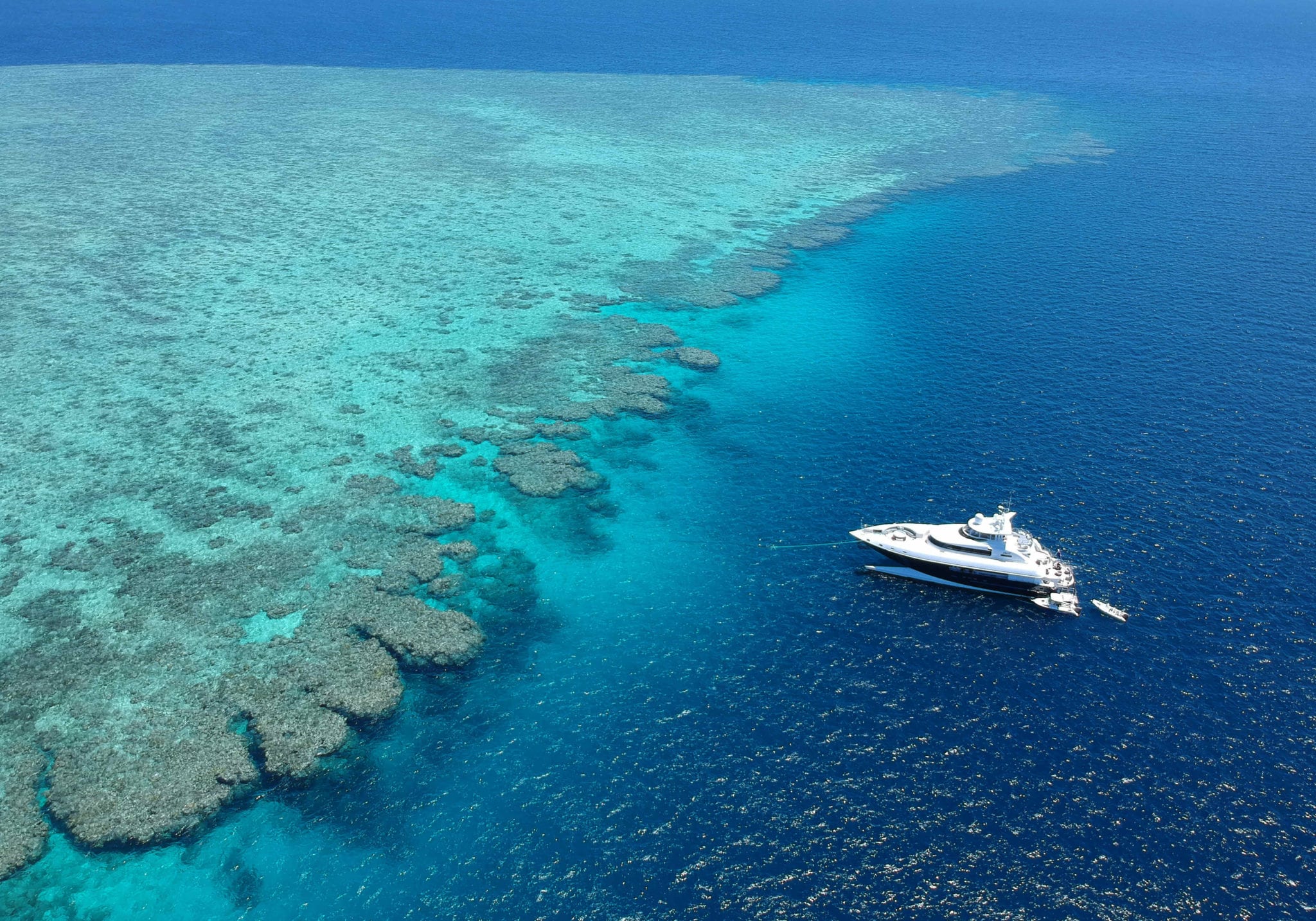 A luxurious yacht anchored near vibrant coral reefs in crystal-clear blue waters. The reef is visible beneath the surface, showcasing various shades of turquoise and green.