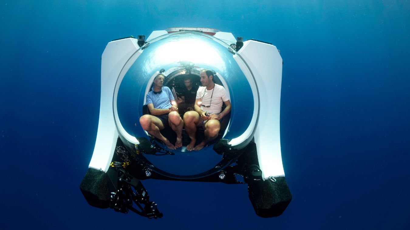 A submarine craft with a large transparent dome contains three people seated inside, surrounded by deep blue water. The atmosphere conveys exploration and adventure in an underwater environment.