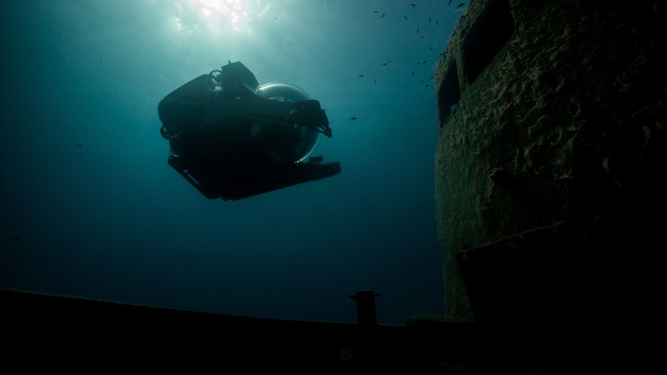 A submersible explores the depths of the ocean near a submerged shipwreck. Sunlight filters through the water, creating a serene atmosphere with small fish swimming around.