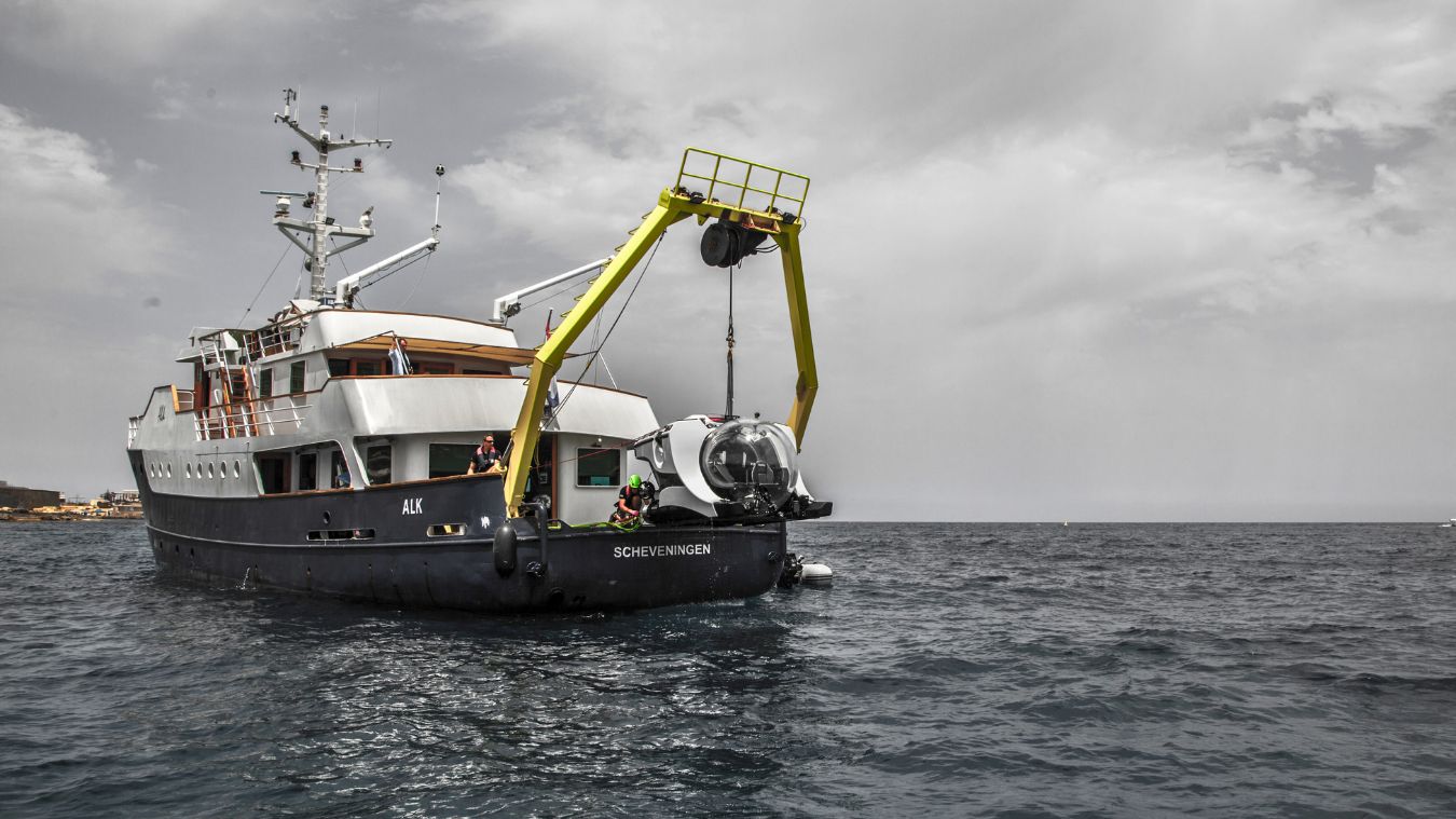 A research vessel with a yellow crane extending over the side is positioned on calm waters. A small submersible is being lowered into the sea, while crew members observe from the deck. The sky is overcast, adding a dramatic backdrop to the scene.