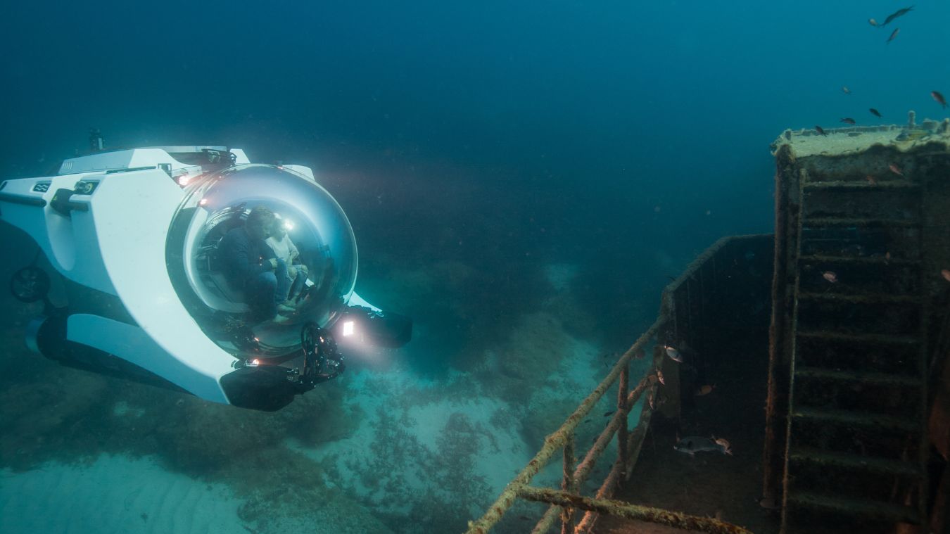 A submersible with a large glass dome exploring the wreck of a ship underwater, surrounded by fish and marine life.