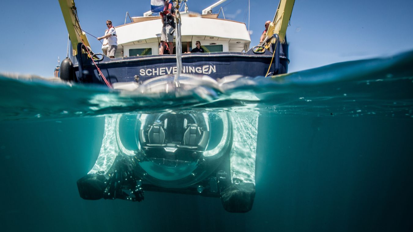 A transparent underwater submersible hovering just beneath the surface, with a research vessel named "Scheveningen" visible above. Several people are on the deck of the vessel. Sunlight filters through the clear water, illuminating the submersible.