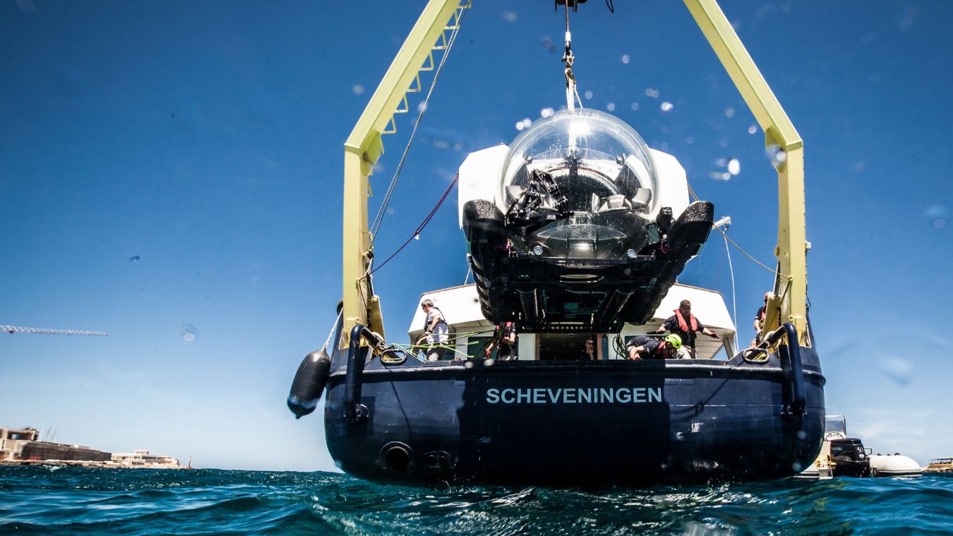 A submersible named Scheveningen is lowered into the water from a ship. The clear blue sky reflects on the surface while crew members prepare for the descent.