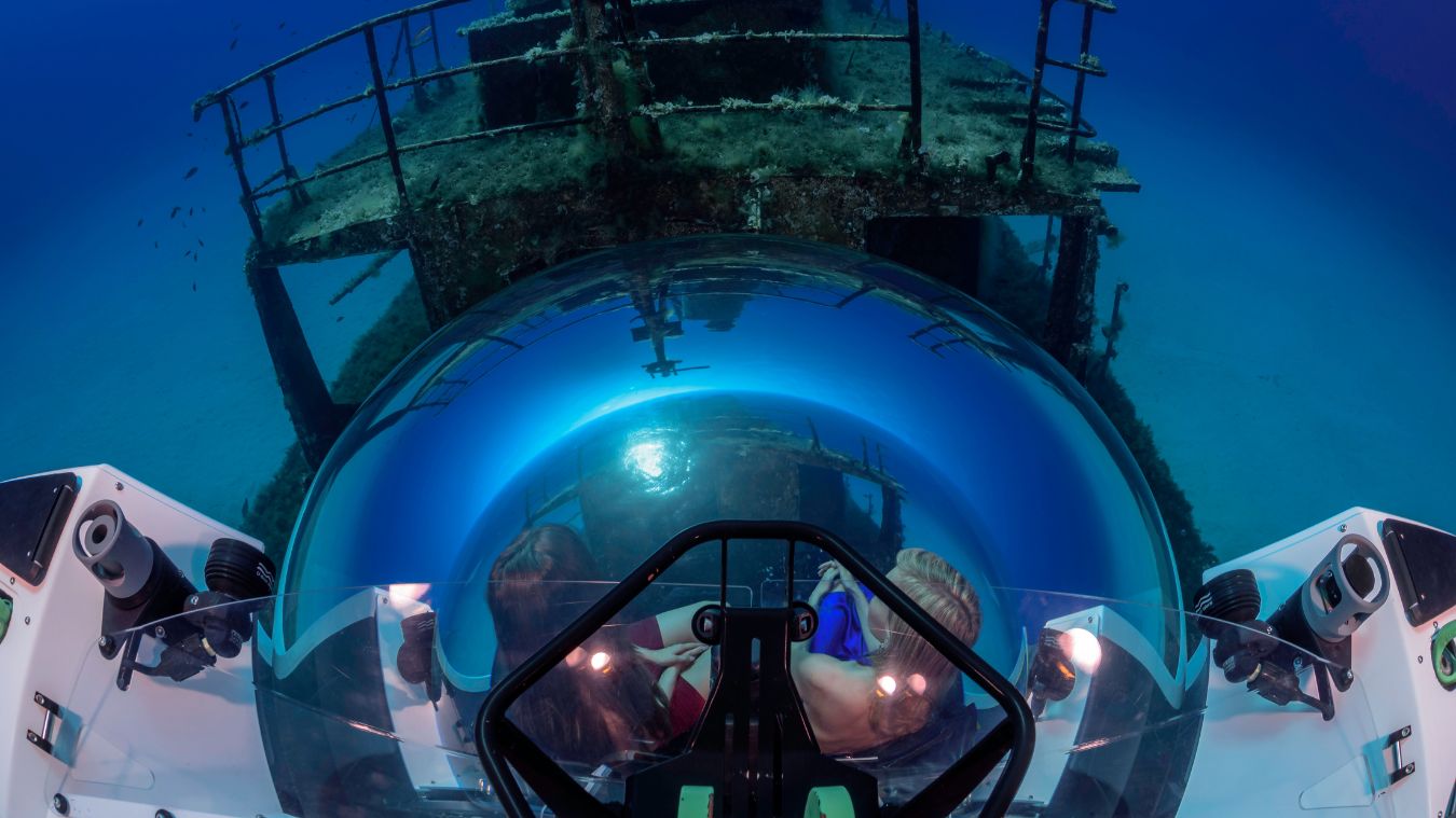 A submarine cockpit view shows two divers exploring a sunken shipwreck beneath clear blue water, with marine life visible nearby.
