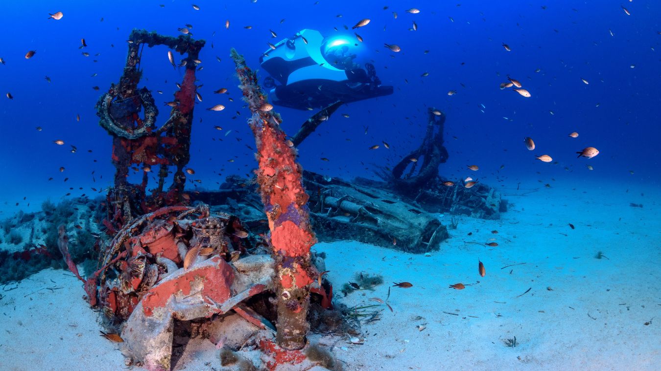 A submerged wreck with bits of metal and coral, surrounded by small fish, under a vibrant blue ocean. A submersible with a bright light explores the site in the background.